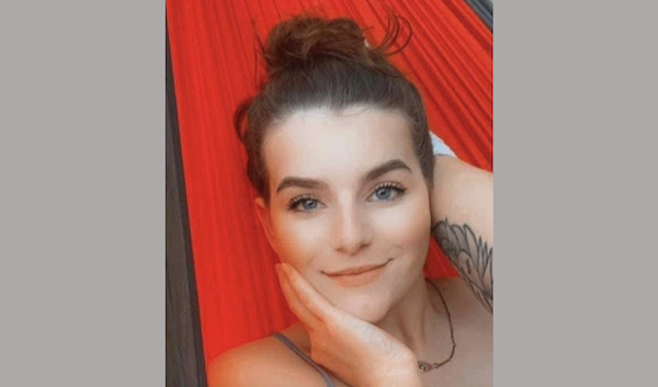 Portrait of a woman with clear skin and dark hair pulled back, holding her chin in her hand while resting on a red hammock, displaying a calm and cheerful expression.