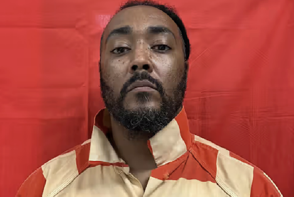 Man in prison attire stands straight against a vivid red background, his shirt collar open, and his expression serious.
