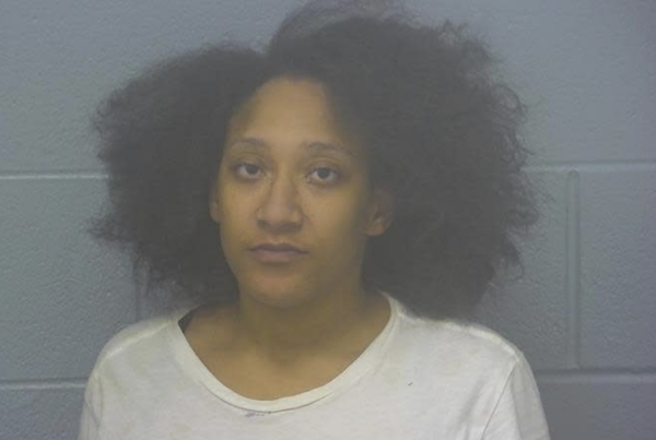 Woman stands for a police booking photo in a light-colored t-shirt, her natural, thick hair framing her face, with a cinderblock wall backdrop.