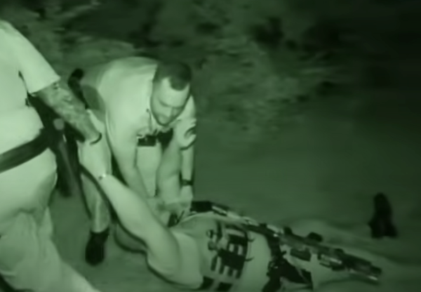 Officers in tactical gear are captured with night vision as they help a fallen deputy, pulling him along a path surrounded by dense foliage.