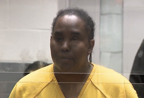 Female defendant in yellow inmate clothing stands for a hearing, separated by a glass divider.