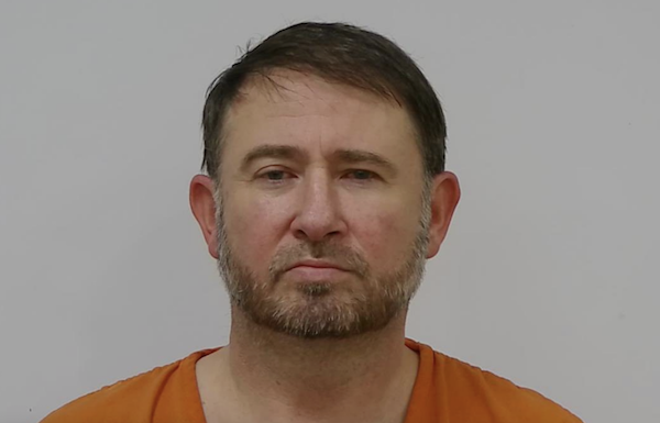 Close-up of an adult male inmate in an orange jumpsuit, looking directly at the camera with a serious expression and a plain background.