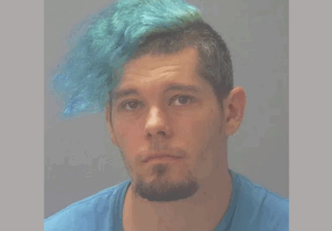 Image showing a person with a side-swept blue hairstyle, wearing a blue t-shirt, with a gray background.