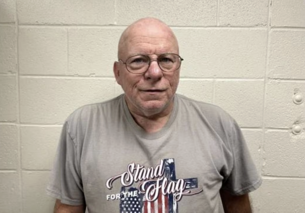 Man with a serious expression, wearing spectacles and an American flag shirt, photographed indoors against a painted concrete wall.