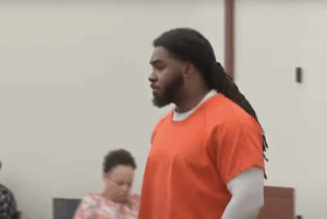 Person in orange prison attire with white undershirt stands solemnly in a courtroom, while another person looks down at a desk in the background.