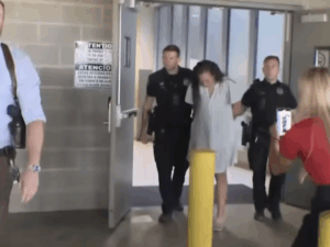 A group exits a building as police officers hold a restrained woman in custody, passing a yellow pole as a bystander records the incident.