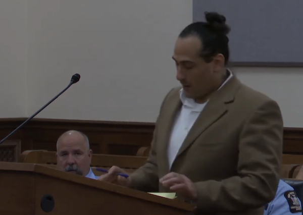 Courtroom scene featuring a speaker in a tan jacket at the podium, with attentive police personnel in the background.