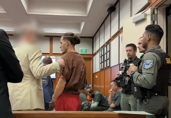 A suspect in a jail outfit faces forward in court, accompanied by his counsel, as uniformed sheriff’s deputies keep watch and other inmates sit along the wall.