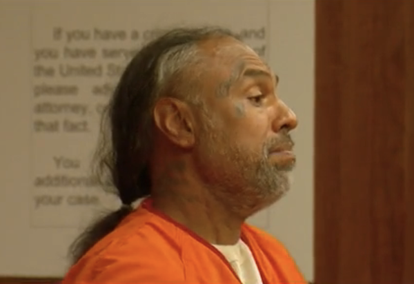 Close-up view of a defendant in an orange jumpsuit during legal proceedings, showing facial tattoos and a solemn expression.