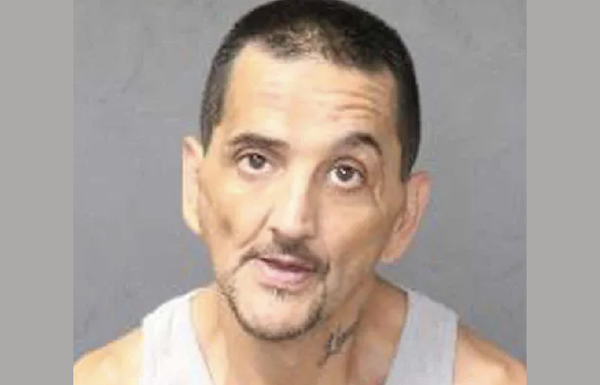 Image of a middle-aged man in a gray tank shirt, appearing unshaven, with a serious expression, photographed against a solid gray background.