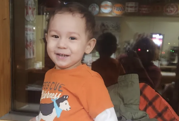 A cheerful little boy poses indoors, his orange shirt featuring a cartoon dog, as a mirror behind him captures both his and an adult’s reflections.