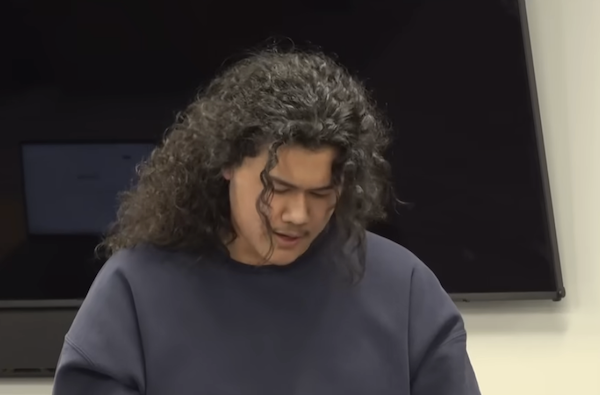 Person with dark, shoulder-length curls reads from a statement, head lowered, during a legal proceeding.