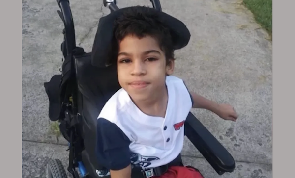 Smiling child with dark hair, wearing a buttoned shirt and red shorts, sits comfortably in a wheelchair on a sunlit path.