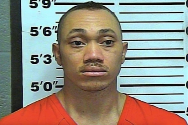 Arrest photo of a person in an orange jumpsuit in front of a police height chart, facing the camera.