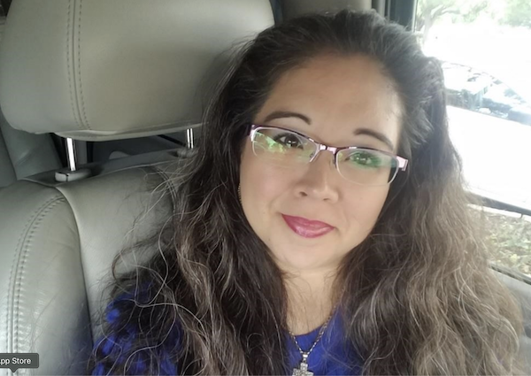 Individual with glasses, long curly hair, and a necklace poses for a selfie in the passenger seat of a car on a bright day.