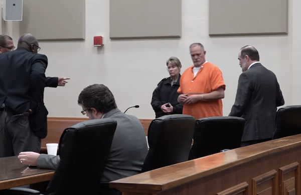In a courthouse, a defendant in orange jail attire stands with hands cuffed, accompanied by a security officer, as lawyers converse nearby and one person is seated at the defense table.