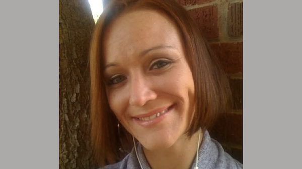 Smiling woman with short, reddish-brown hair poses outdoors near a brick wall and tree trunk.