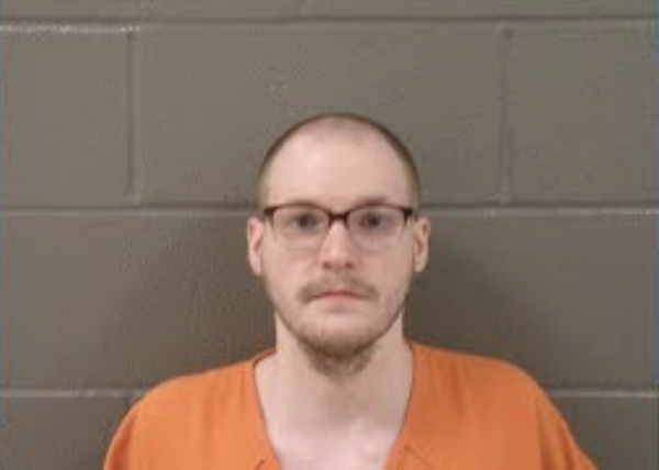 Portrait of a man in correctional clothing and glasses, facing the camera straight-on, positioned before a gray institutional wall.