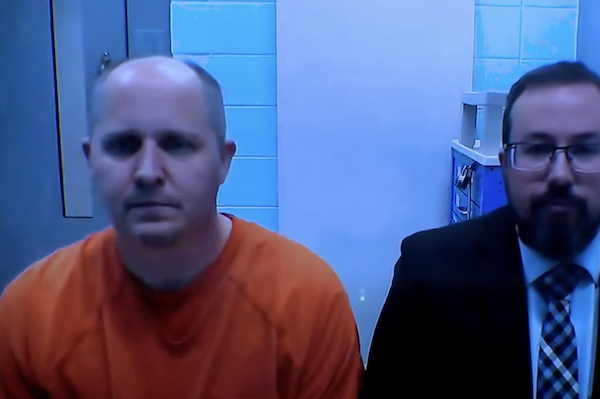 A person in orange prison clothing is joined by a man in formal business attire, both facing forward on a video feed with light blue and white tiles behind them.