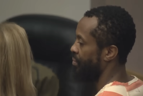 A close-up image of a man in a jail uniform attending a legal proceeding, with another person partially visible in the foreground.