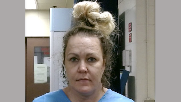 A close-up photo of a woman with a tired or distressed look, wearing a light blue garment, her blond hair tied up, posed in front of an off-white wall and office-like environment.