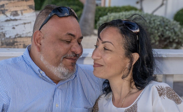 A man and woman look at each other and smile, sitting side by side against a white fence with leafy bushes and a tree behind them.