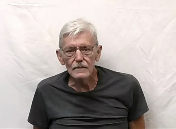 Portrait of a gray-haired man in glasses and a dark shirt, positioned against a solid white wall.