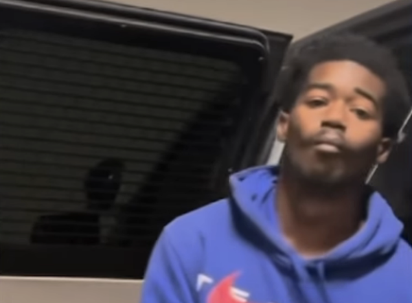 Individual in a blue hoodie poses in a dimly lit indoor setting, with a dark vehicle window and metallic surfaces in the background.