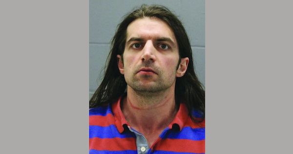 Portrait-style booking image of an adult male with straight long hair, appearing in a striped shirt in front of a plain gray backdrop.