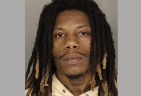 Police booking photo of a man with tan complexion and dark-and-blonde dreadlocks, showing little emotion, photographed from the shoulders up.