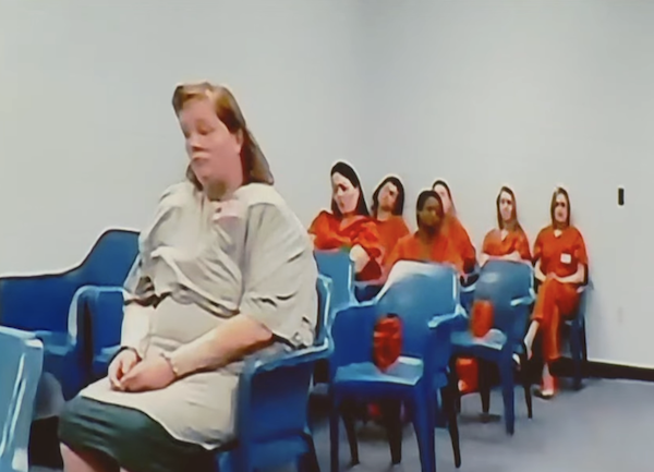 In a room with plastic chairs, a group of women in prison attire are seated in a staggered formation, with the focus on a woman in light-colored clothing sitting closest to the camera.