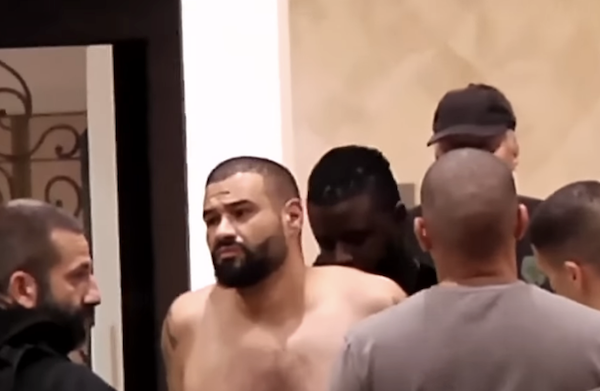 In an indoor hallway, officers escort a shirtless man with short hair and a beard, his arms behind his back as they prepare to move him.