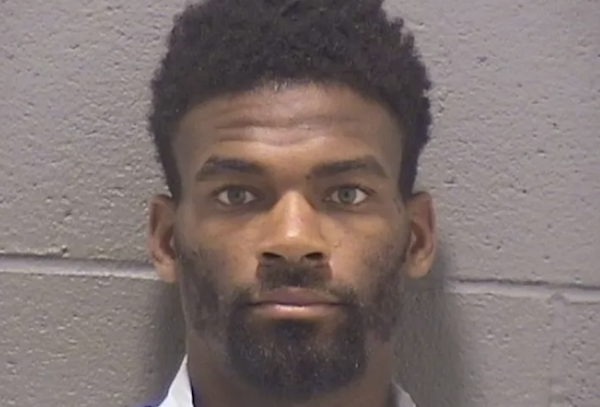 Police custody photo of a man in a white shirt, standing against a gray brick wall, looking directly at the camera.