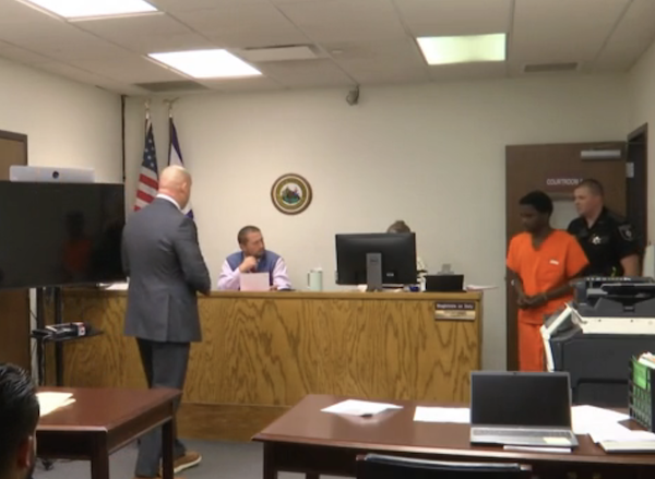 The judge's bench is the focal point as attorneys, law enforcement, and a detainee in orange participate in a hearing, with computers and legal documents visible throughout the room.