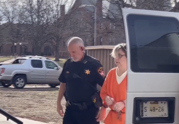 A woman wearing handcuffs and an orange jumpsuit is accompanied by a sheriff’s deputy, exiting a transport van in a parking area with trees and buildings behind them.