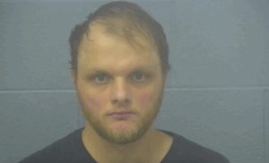 Booking image of a bearded man with receding hair, unsmiling, standing before a plain concrete wall, lit with subdued lighting.