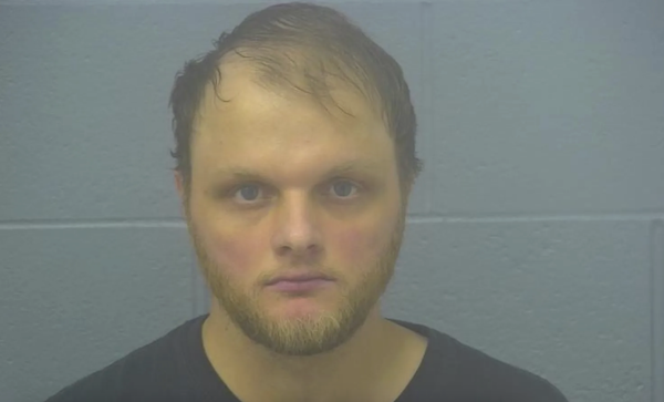 Booking image of a bearded man with receding hair, unsmiling, standing before a plain concrete wall, lit with subdued lighting.