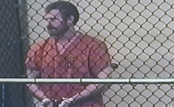 Detainee in an orange uniform with handcuffs is photographed behind a metal fence inside a holding area.