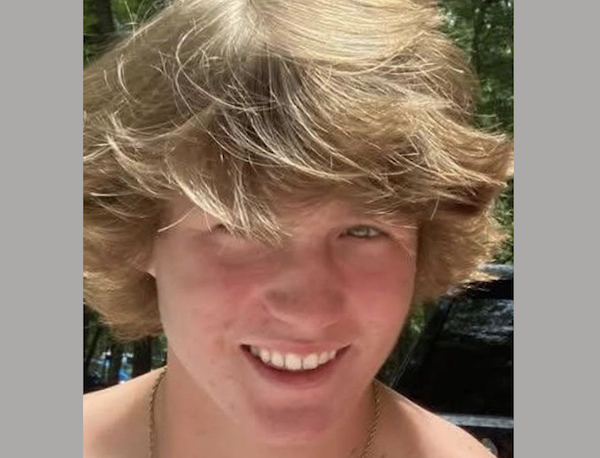 Teen boy in natural light, grinning widely, his long hair partially covering his forehead, green leafy scenery behind him.