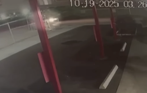 Security image features a dark parking lot with a vehicle’s headlights illuminating the sidewalk, red support beams visible in the foreground, and white lot markings nearby.