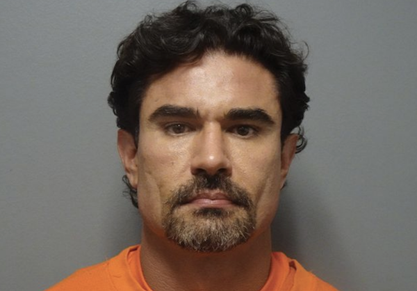 Portrait-style photo showing a man with a stern expression, dark facial hair, and curly hair, dressed in orange with a gray backdrop.