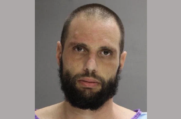 A close-up photograph of a man with short brown hair and a dark beard, dressed in a blue shirt, with a blank gray background behind him.