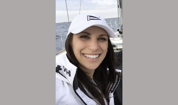 Close-up selfie of a woman outdoors on a boat, dressed in a white Helly Hansen jacket and white hat, with water and part of the sailboat in the background.