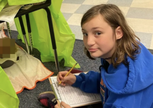 A school-aged child works on paperwork during an indoor activity, positioned next to a table transformed into a small shelter with a fluorescent covering; an unidentified, blurred item is underneath the desk.