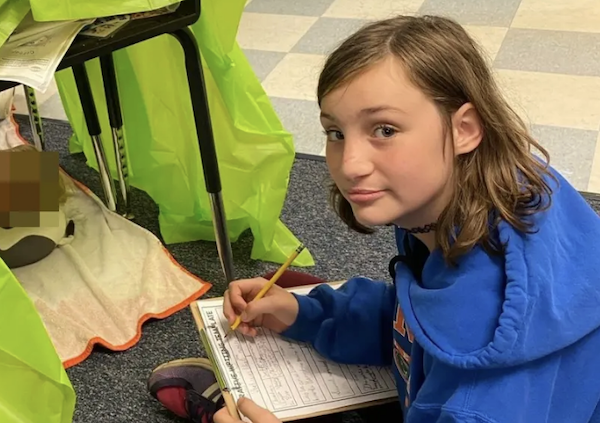A school-aged child works on paperwork during an indoor activity, positioned next to a table transformed into a small shelter with a fluorescent covering; an unidentified, blurred item is underneath the desk.
