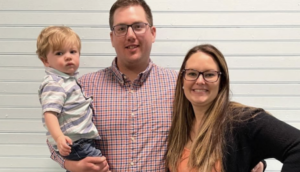 A young family with a father holding his son and a mother standing close by, all facing the camera against a paneled wall background.