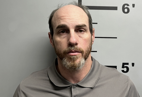 Bearded man in a gray shirt poses for a police booking photo with a neutral look, standing in front of a height chart on the wall.