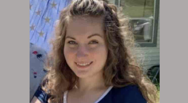 Close-up image of a woman with light brown curly hair and a friendly expression, standing in a yard near a window and patterned fabric.