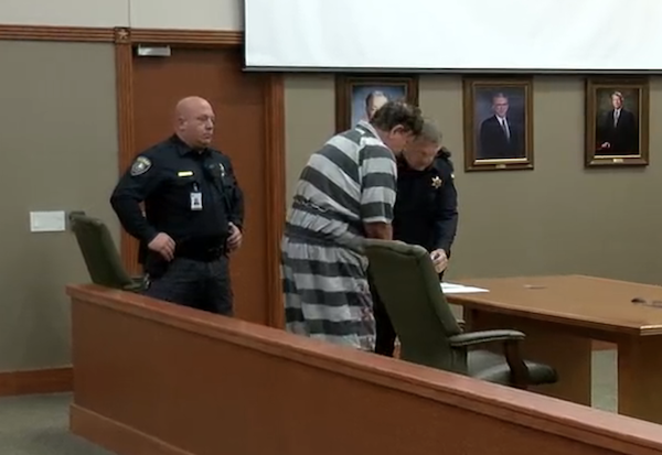 A detainee in jail uniform stands before a judge’s bench, watched by a security officer, with courtroom portraits of legal figures visible on the wall.