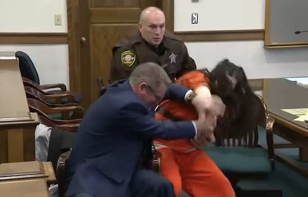 A tense moment unfolds in court as a law enforcement officer and a suited man jointly restrain an agitated individual in prison clothing seated at a legal table.